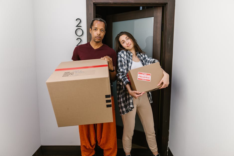 A young woman with long brown hair smiling at the camera while holding a large empty cardboard box labeled 'Fragile Glass' on the side, inside a room with a textured blue wall. Around her, there are s