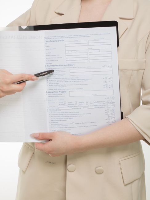 A professional woman with long black hair, dressed in a grey vest and light-colored blouse, stands indoors holding a clipboard labeled 'Home Insurance Policy' and a pen, while explaining policy detail